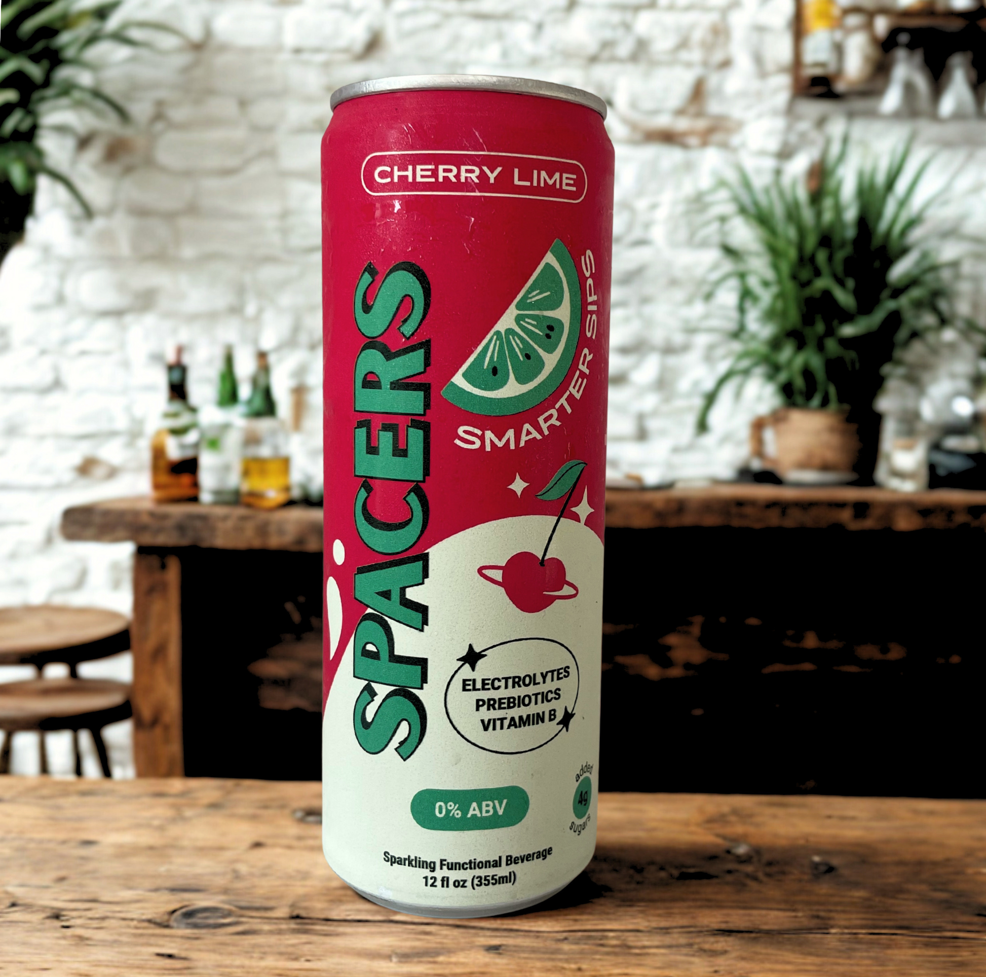 Spacers Cherry Lime 12 oz slim can displayed on a rustic wooden bar with a white brick wall and green plants in the background. The vibrant red and mint can features “0% ABV” and highlights electrolytes, prebiotics, and vitamin B—perfect for health-conscious and sober curious consumers seeking a functional, non-alcoholic drink.